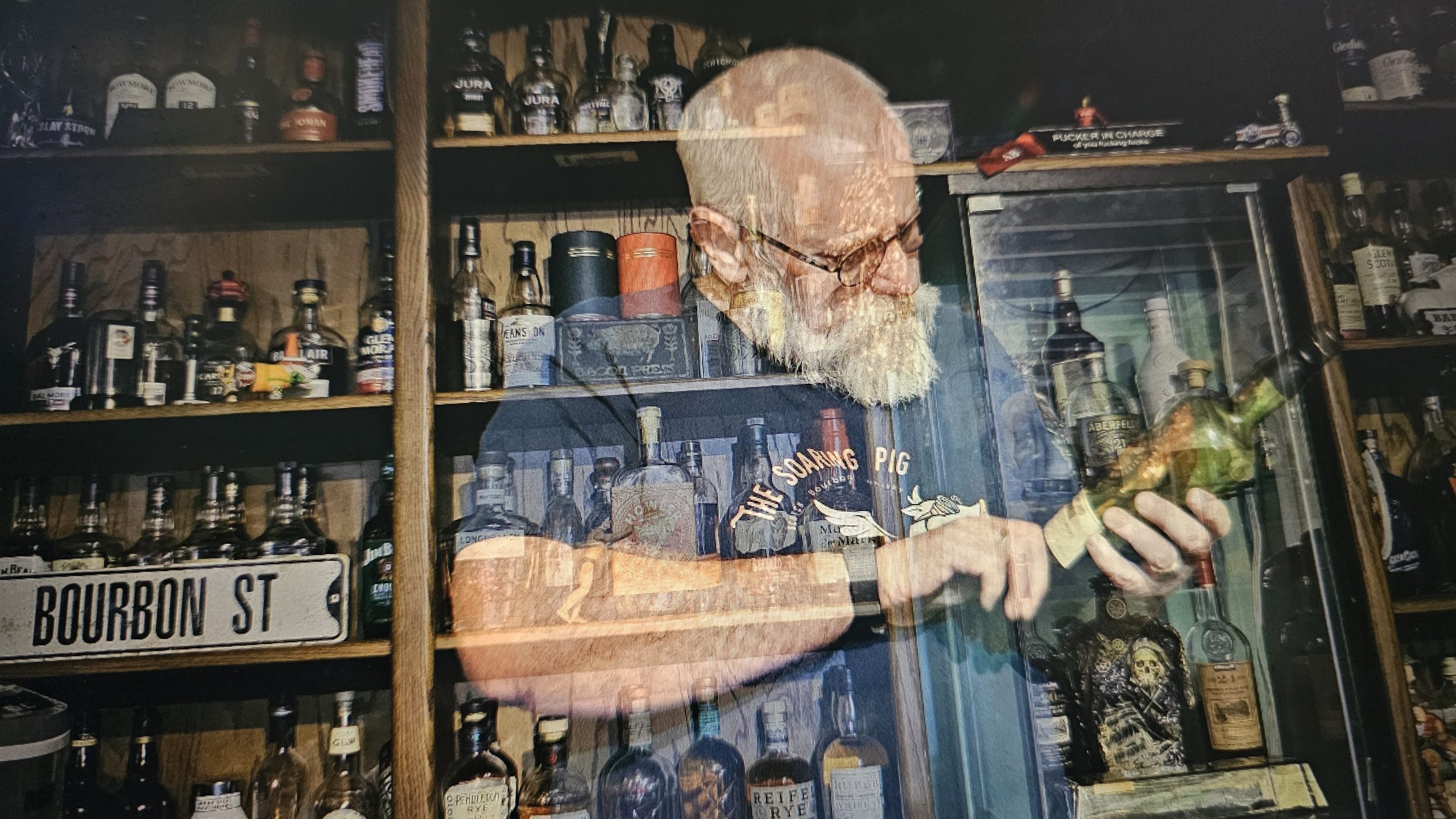 Stephen Dafoe in his Soaring Pig whiskey bar, examining a bottle, surrounded by an extensive whiskey collection. Unique double exposure effect adds depth.