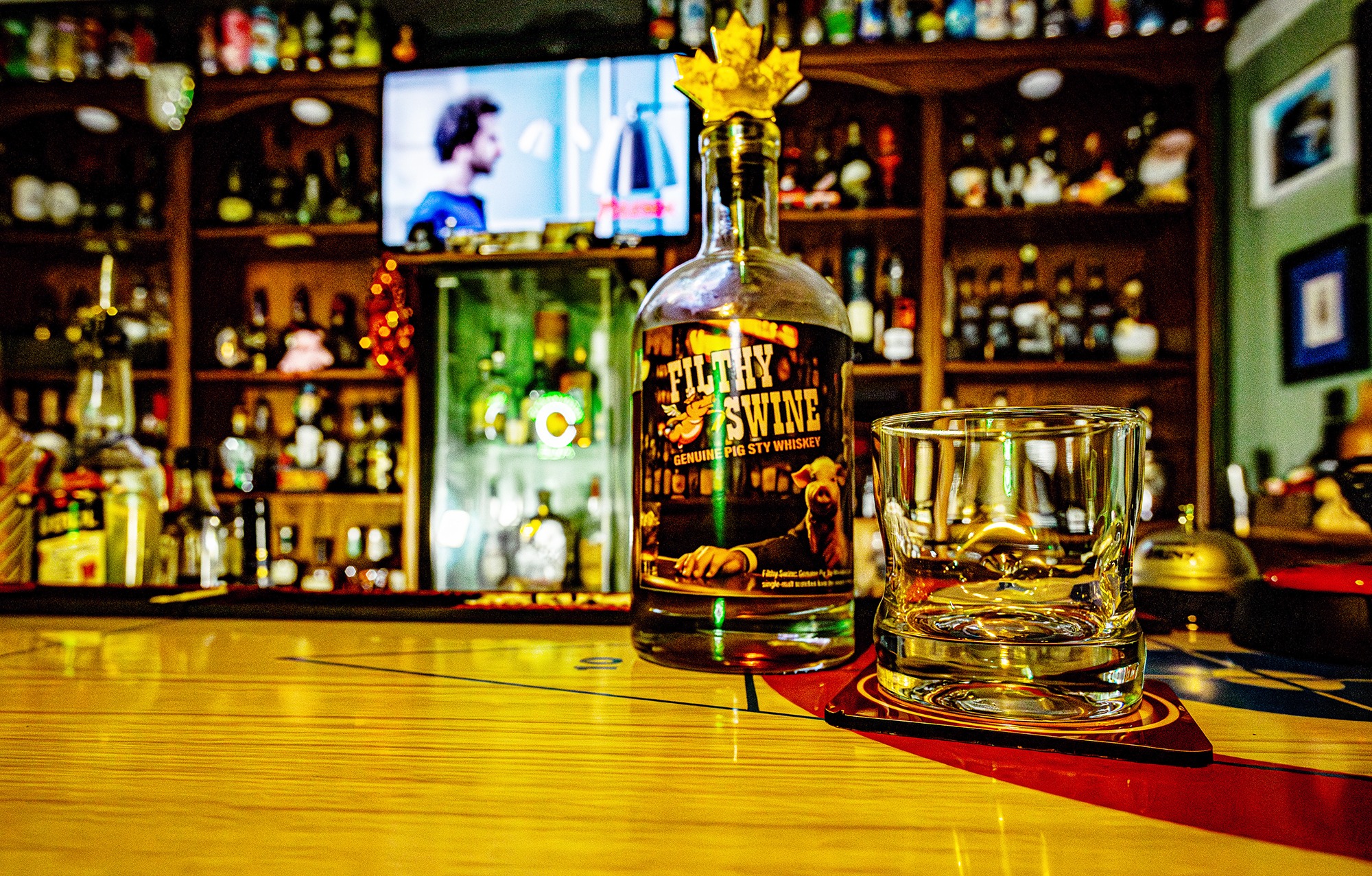 A bottle of Filthy Swine Genuine Pig Sty Whiskey with a whiskey glass on the bar counter at The Soaring Pig Bar, surrounded by an extensive whiskey collection.